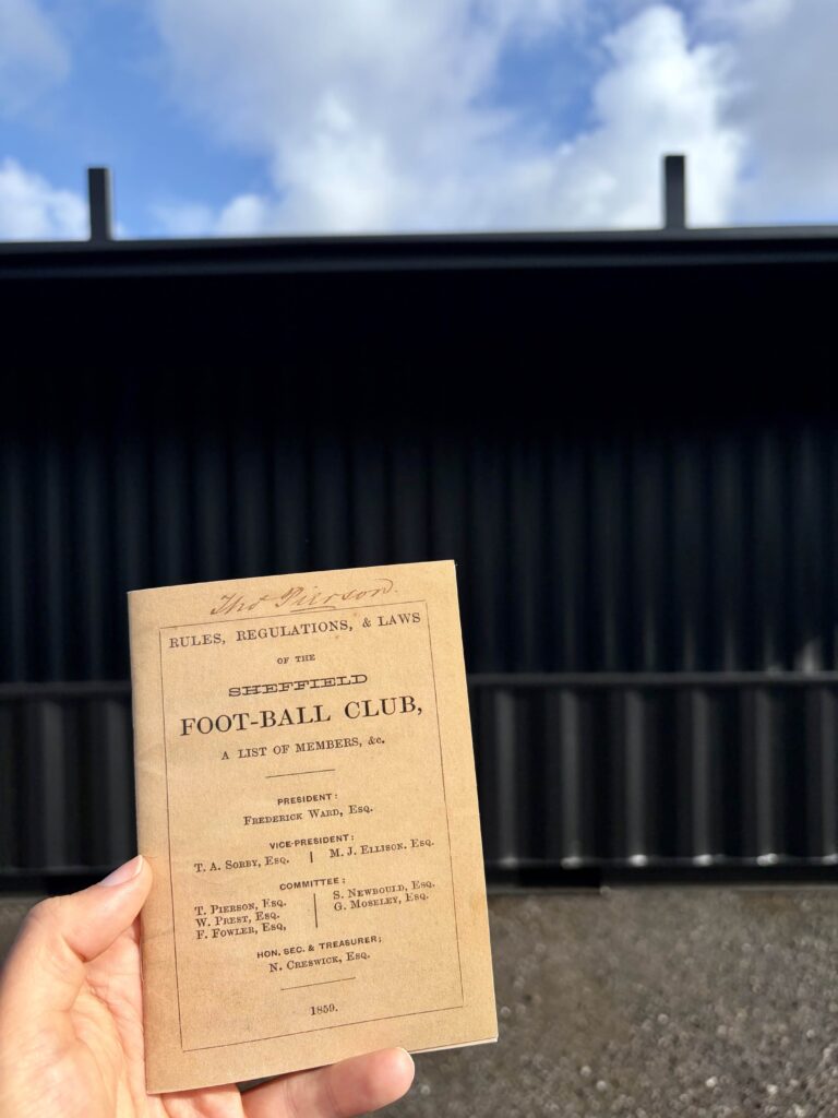 First football rule book from Sheffield Football Club with shipping container stadium stands in the background.